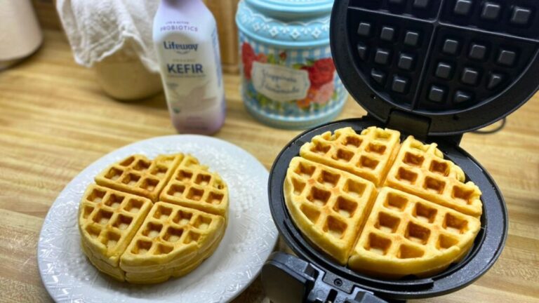 Golden homemade waffles stacked on a plate beside a waffle iron on a wooden kitchen counter