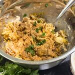 Mixing bowl filled with seasoned shredded chicken, herbs, and cheese on a dark wood table