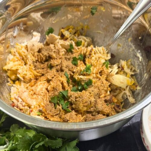 Mixing bowl filled with seasoned shredded chicken, herbs, and cheese on a dark wood table