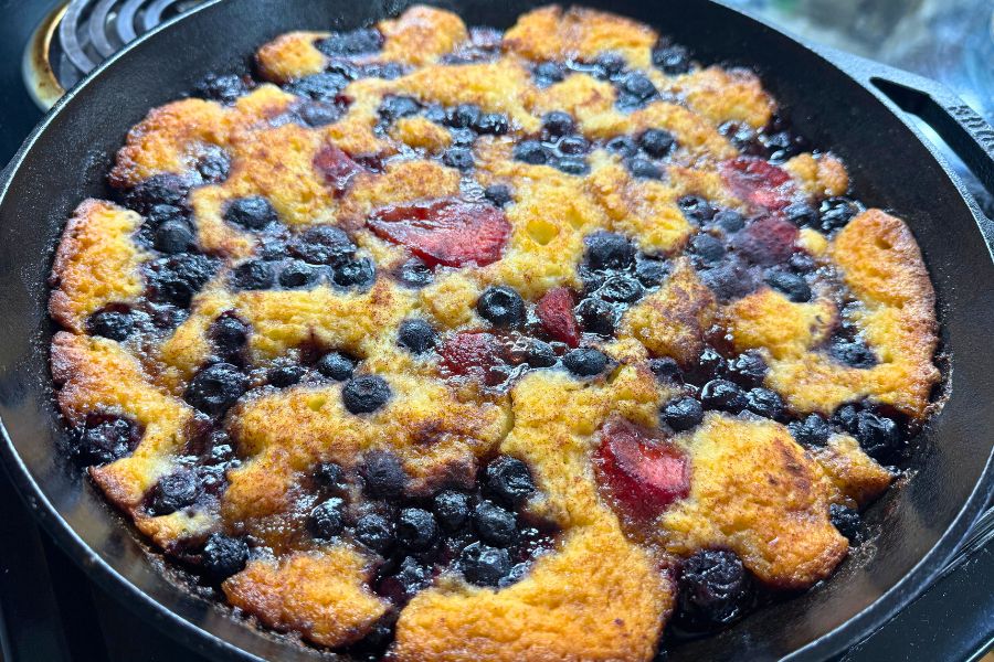 Golden sourdough berry cobbler bubbling in a cast iron skillet, filled with blueberries and strawberries.