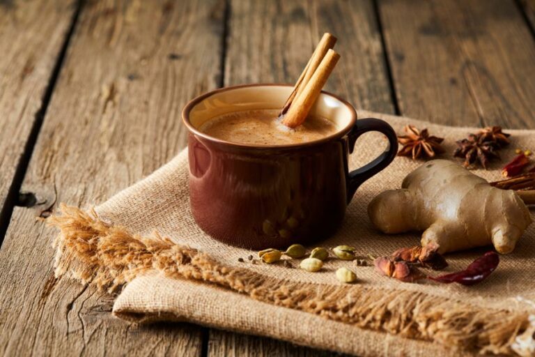 A rustic brown mug of spiced latte with cinnamon sticks, surrounded by fall spices like ginger, cardamom, and star anise on a burlap cloth over wooden planks.