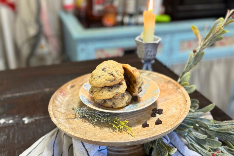 Stack of golden sourdough chocolate chip cookies on a floral plate, styled on a wooden cake stand with chocolate chips and greenery, set beside a glowing candle.