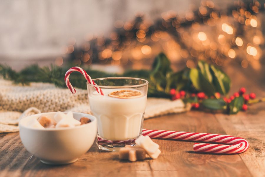 Glass of creamy white drink with a candy cane, surrounded by caramels, evergreen branches, and twinkle lights, styled for a cozy Christmas setting.