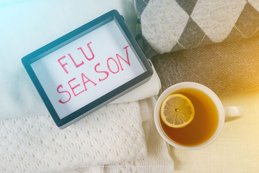 Cup of tea with lemon and a sign reading “Flu Season” on cozy knit blankets and sweaters.