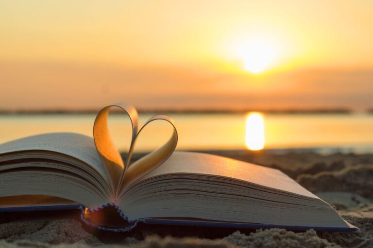 An open book with pages shaped into a heart, resting on sand at sunset near a calm body of water.