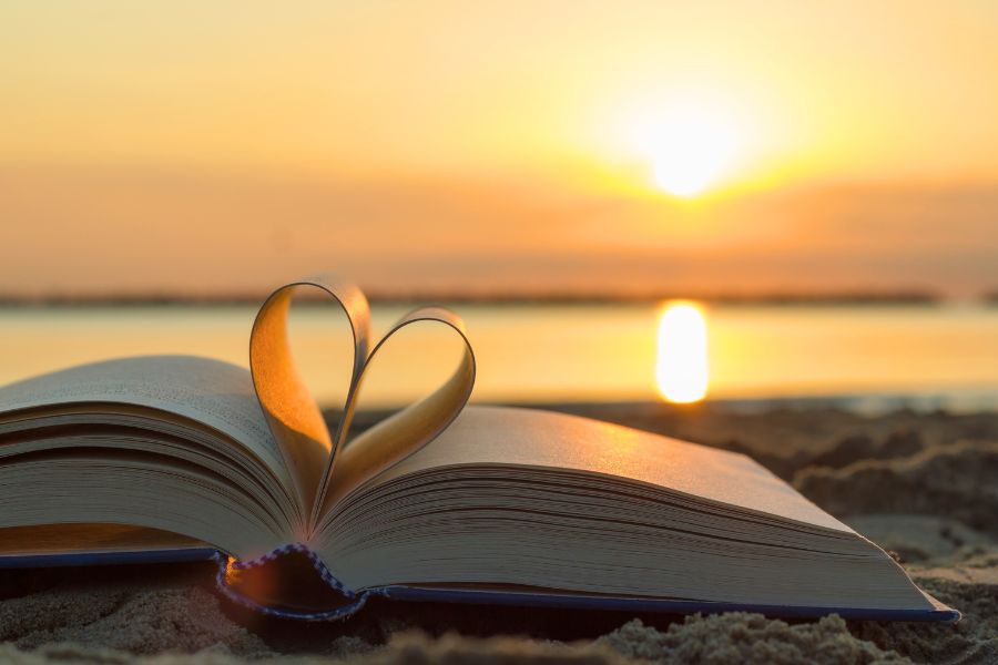 An open book with pages shaped into a heart, resting on sand at sunset near a calm body of water.