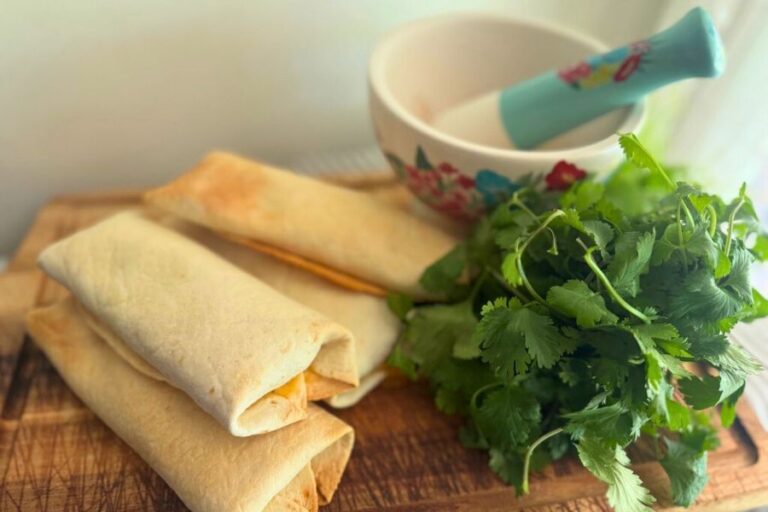 Baked chicken taquitos on a wooden board with fresh cilantro and a floral mortar and pestle