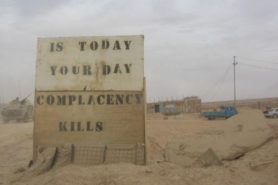 A large weathered military sign in a desert reading “IS TODAY YOUR DAY – COMPLACENCY KILLS”