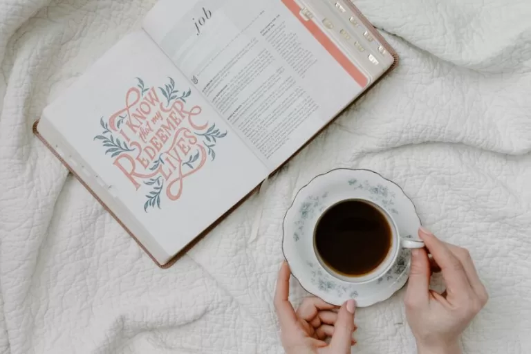 Open Bible showing the book of Job and hand holding a floral teacup on a cozy white blanket.