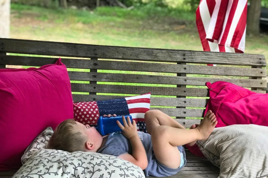 Child resting on a porch swing with patriotic pillows and an American flag in the background, symbolizing a peaceful, meaningful 4th of July.