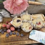 Sourdough blueberry and strawberry muffins on a wooden board surrounded with florals and strawberries and blueberries.