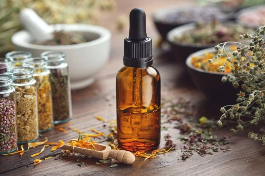 Amber essential oil bottle surrounded by dried herbs, glass jars, and a wooden scoop on a rustic table