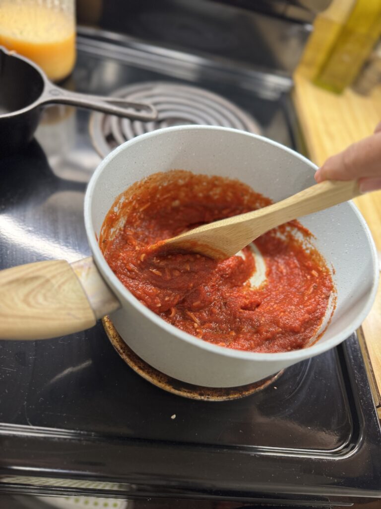 Homemade pizza sauce simmering in a saucepan with a wooden spoon on the stovetop.
