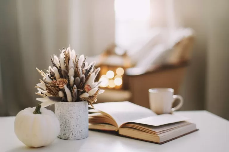 A cozy fall scene with a white pumpkin, dried flower bouquet, open book, and coffee mug on a white table, softly lit by morning sunlight.