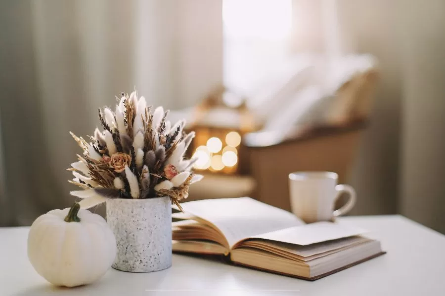 A cozy fall scene with a white pumpkin, dried flower bouquet, open book, and coffee mug on a white table, softly lit by morning sunlight.