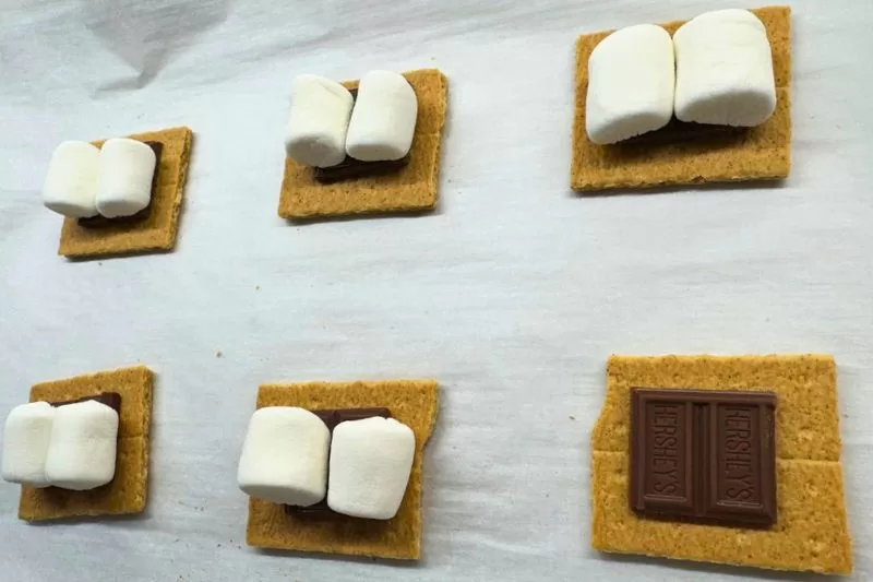 Graham crackers topped with Hershey’s chocolate and marshmallow halves on parchment paper, showing the base layer for s’mores chocolate chip cookies.