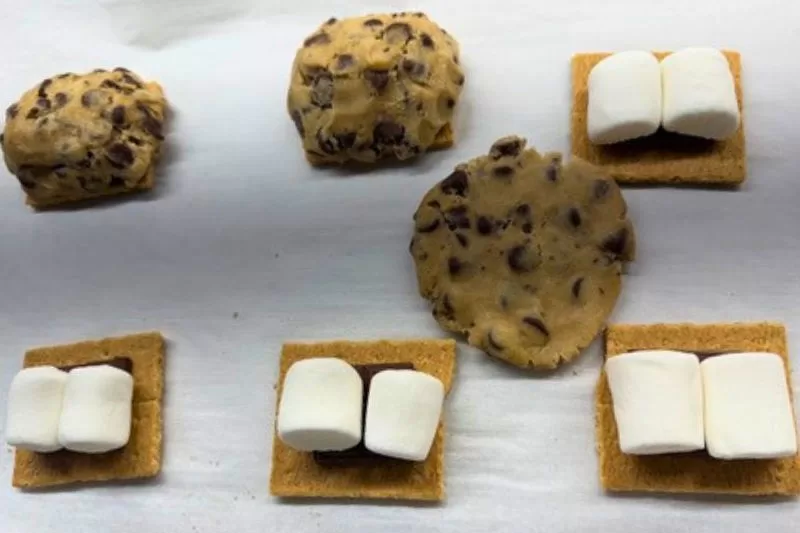Chocolate chip cookie dough mounds beside graham crackers topped with chocolate and marshmallows, showing how to assemble s’mores chocolate chip cookies before baking.