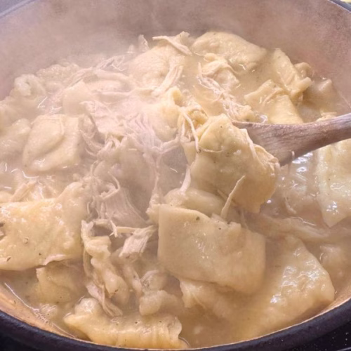 Homemade chicken and dumplings cooking in a Dutch oven with tender shredded chicken and soft dumplings lifted on a wooden spoon.