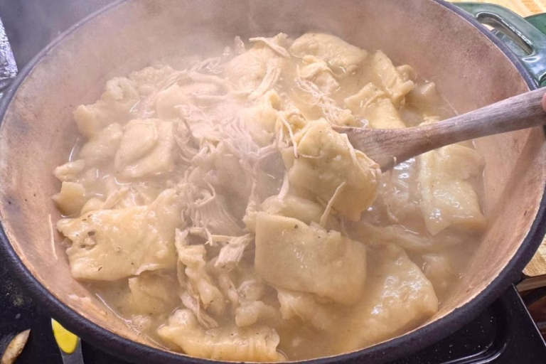 Homemade chicken and dumplings cooking in a Dutch oven with tender shredded chicken and soft dumplings lifted on a wooden spoon.