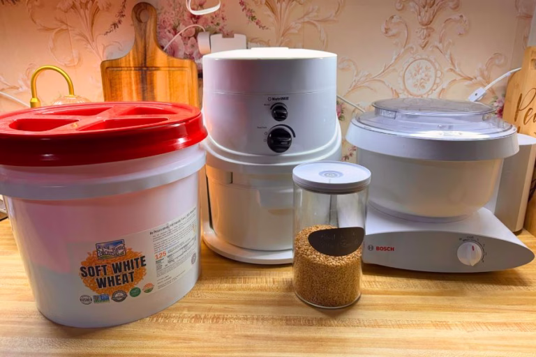 Fresh milled flour tools arranged on a kitchen counter including a NutriMill Classic grain mill, Bosch mixer, wheat berries, and storage bucket used for home baking.