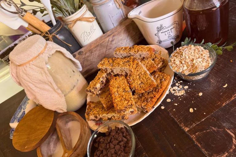 Sourdough granola bars made with sourdough discard, oats, honey, peanut butter, and chocolate chips arranged with ingredients on a farmhouse kitchen table.