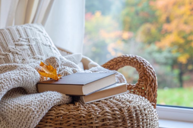 Cozy reading nook with a knitted blanket, books, and autumn leaves outside the window, representing fall essential oil blends for a cozy home.